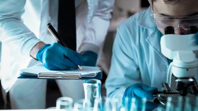 Ein Wissenschaftler arbeitet konzentriert an einem Mikroskop, während ein Kollege mit blauen Handschuhen Notizen auf einem Klemmbrett macht. Im Vordergrund sind Laborutensilien zu sehen.