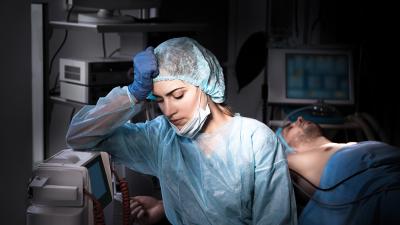 Ärztinn im Op mit traurigem Gesichtausdurck und Faust auf der Stirn, im Hintergurnd ein Mann auf einer OP-Liege.