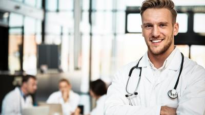 Links vorne ein lächelnder junger Arzt, hinten recht Gruppe Ärzte sitzt um Tisch