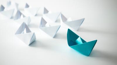 Mehrere weiße Papierboote und ein einzelnes blaues Papierboot auf weißem Hintergrund, symbolisch für Individualität und Führung.