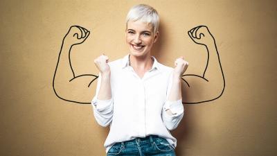 eine blonde Frau steht mit geballten Fäusten vor einer Wand mit umriss von muskolösen Oberarmen