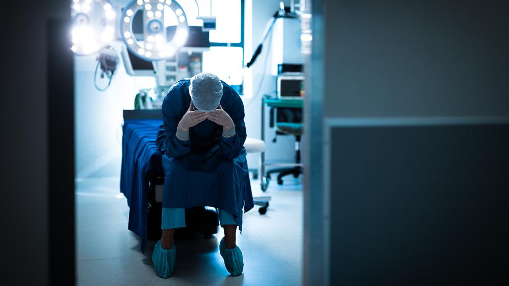 Erschöpfte OP-Kraft in blauer Schutzkleidung sitzt mit gesenktem Kopf in einem Operationssaal.