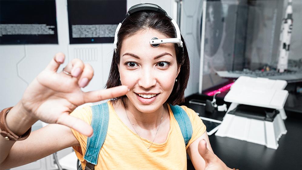 Frau in gelbem Shirt und Brain-Computer-Interface formt mit ihren Händen einen Rahmen, im Hintergrund technische Modelle und Ausstellungsstücke.