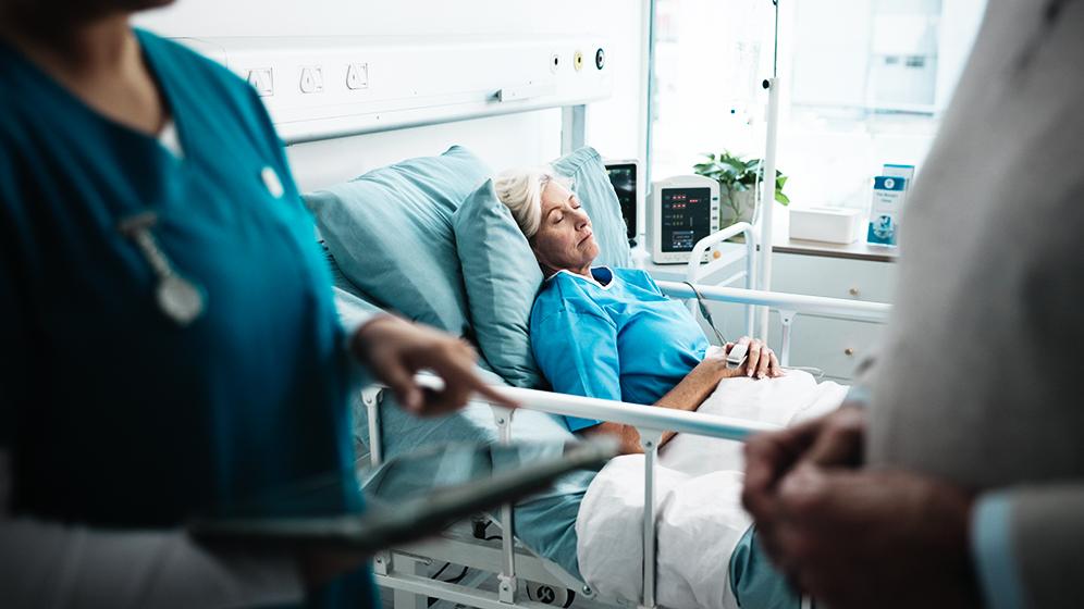 Ältere Patientin liegt schlafend in einem Krankenhausbett, im Vordergrund stehen medizinische Fachkräfte.