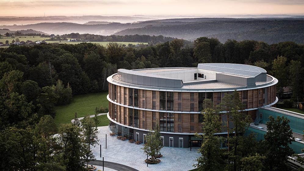 Waldkliniken Eisenberg: Rundes Klinikgebäude mit Fassade aus Holz, im Hintergrund idyllische Hügel im Nebel  